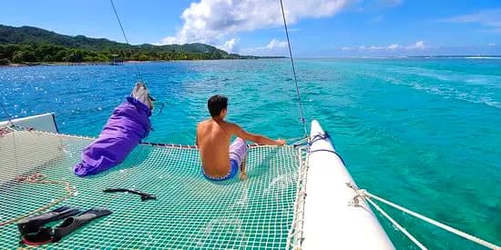 Man sitting on the catamaran's trampoline looking across turquoise water to the green Roatan coast