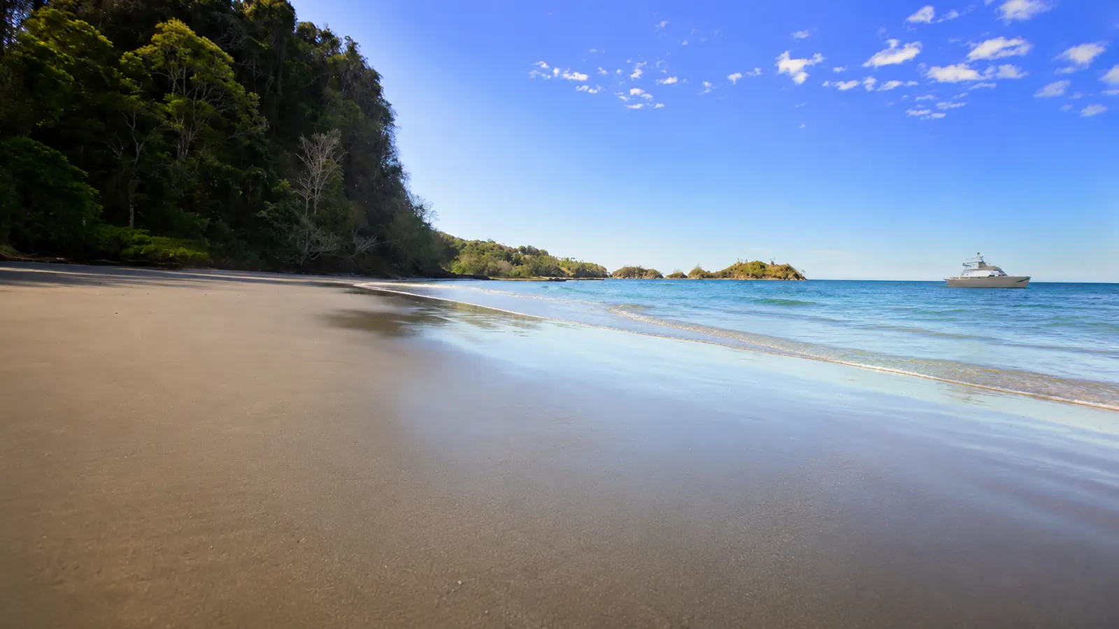 Smooth wet sand of a quiet Tortuga Island beach below forested hills, a boat anchored in the distance