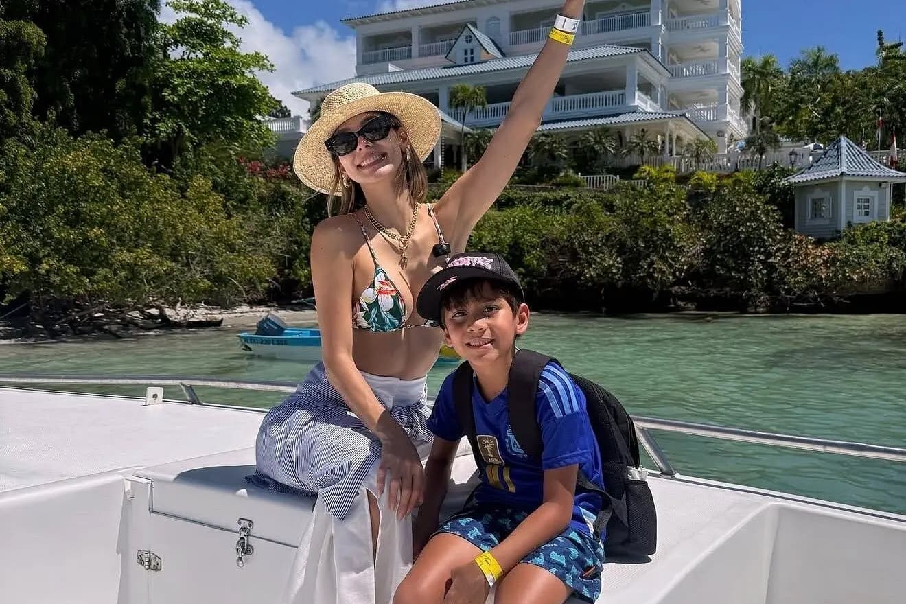 Woman in a sun hat waving beside a boy on the boat's bow, a hillside hotel on the Samaná shore behind