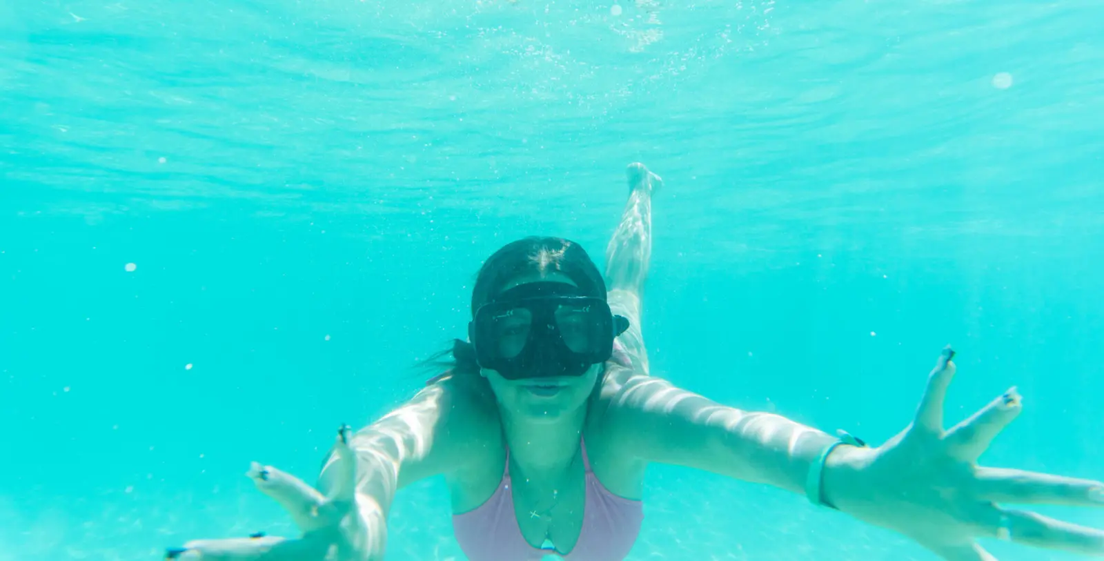 Woman in a snorkel mask swimming underwater toward the camera with arms spread wide
