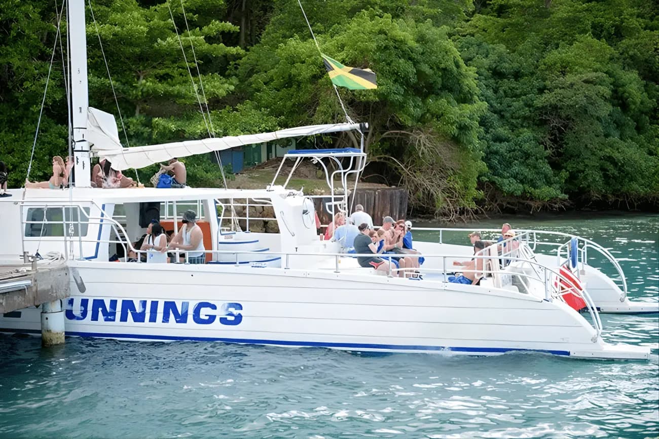 Guests aboard a white catamaran with a Jamaican flag, tied up at a dock by a leafy shore