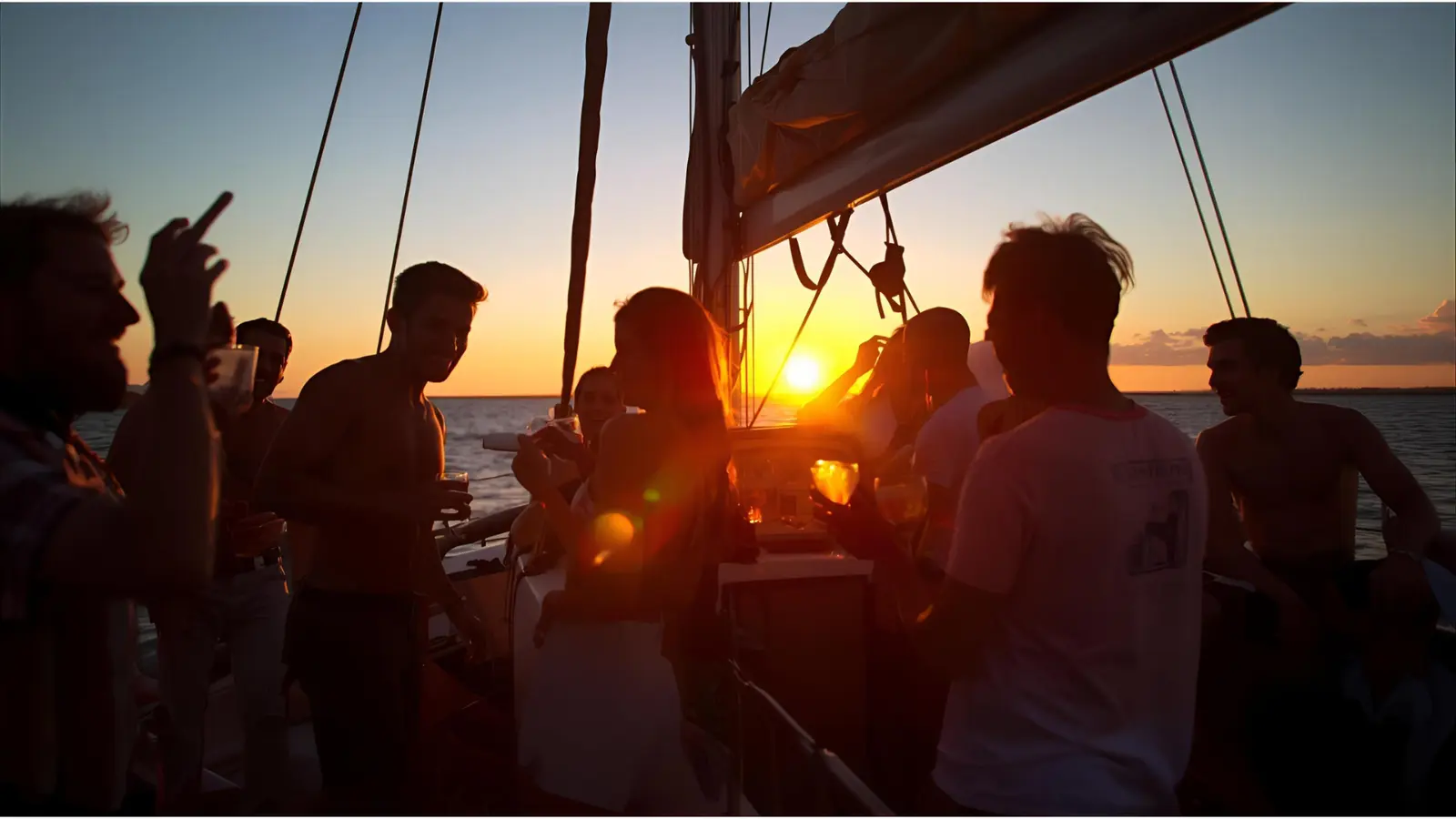 Silhouetted guests with drinks under a sailboat's boom as the sun meets the horizon on Bacalar Lagoon