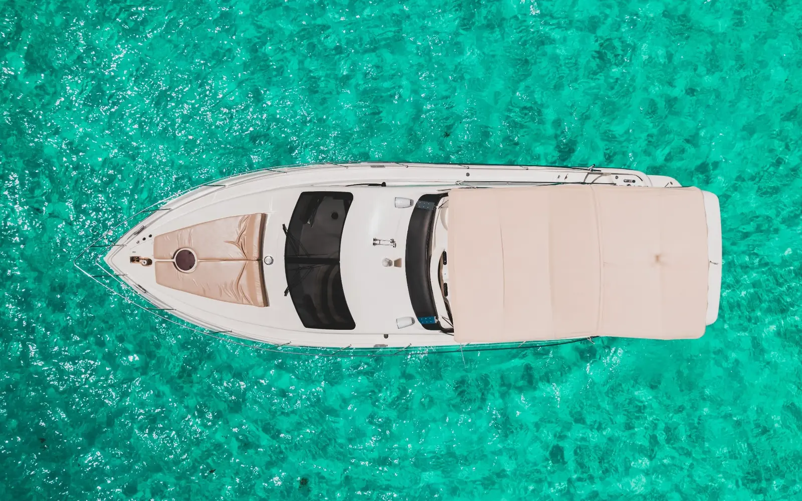 Top-down drone shot of a white motor yacht with a tan bow sun pad and cream canopy on turquoise water