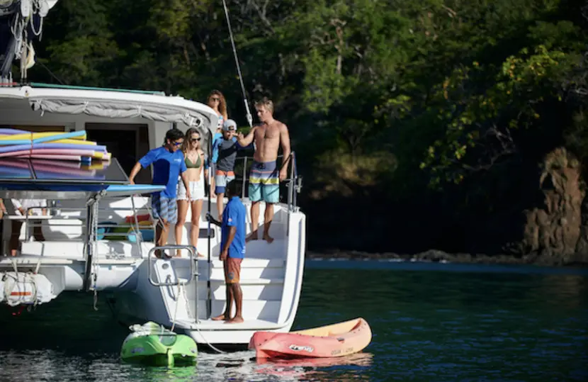 Guests on the catamaran's stern steps above two kayaks, paddleboards stacked on deck by a forested shore