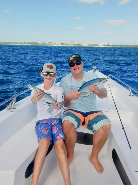 Boy and man seated on the bow, each holding a small fish, deep blue sea behind