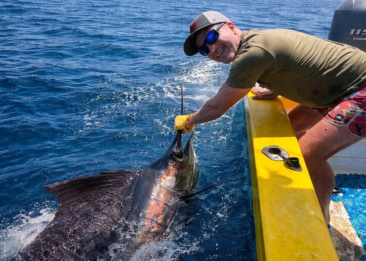 A man in gloves reaches over the panga's yellow gunwale to grip a sailfish at the surface