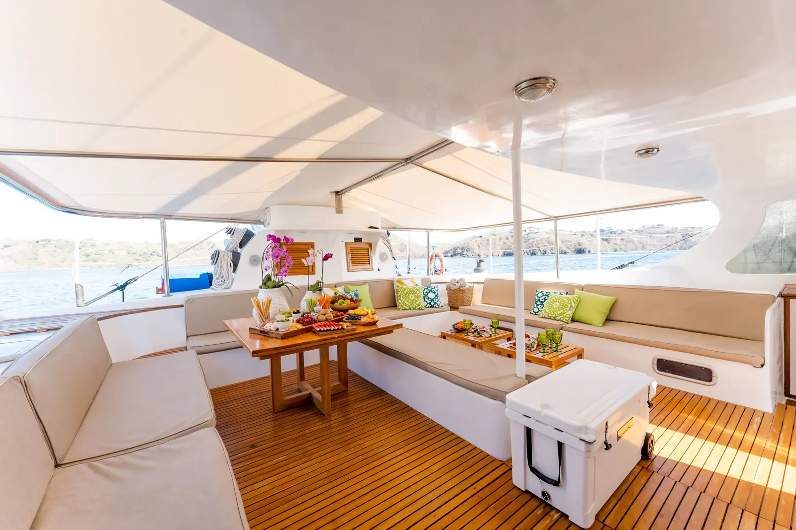 Shaded teak aft deck of the catamaran with cushioned seating, snack tables and a cooler