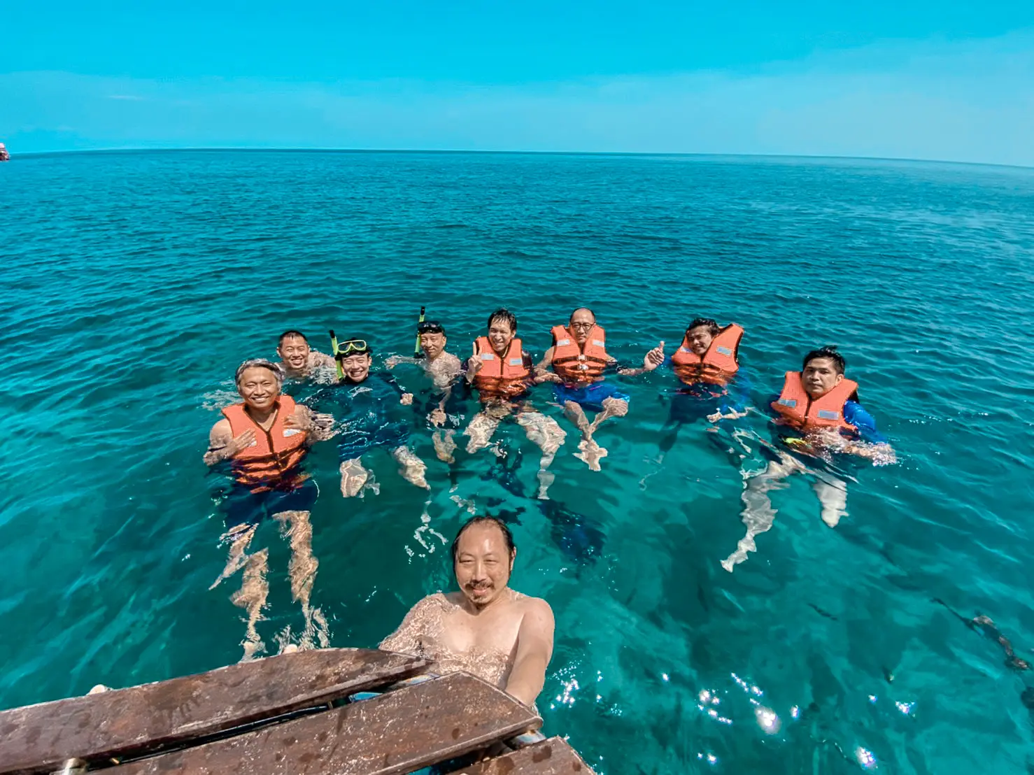 Guests in orange life jackets floating in turquoise water behind a smiling man taking a selfie
