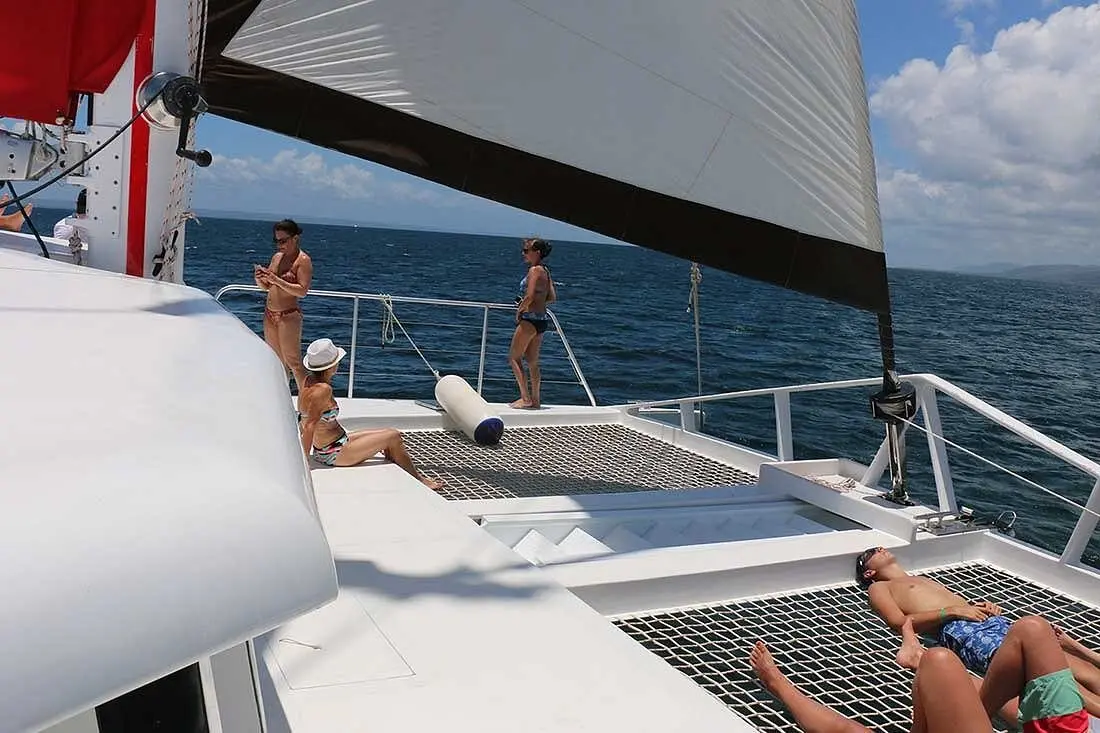 Guests standing at the bow rail and sunbathing on the nets of a sailing catamaran under a white sail