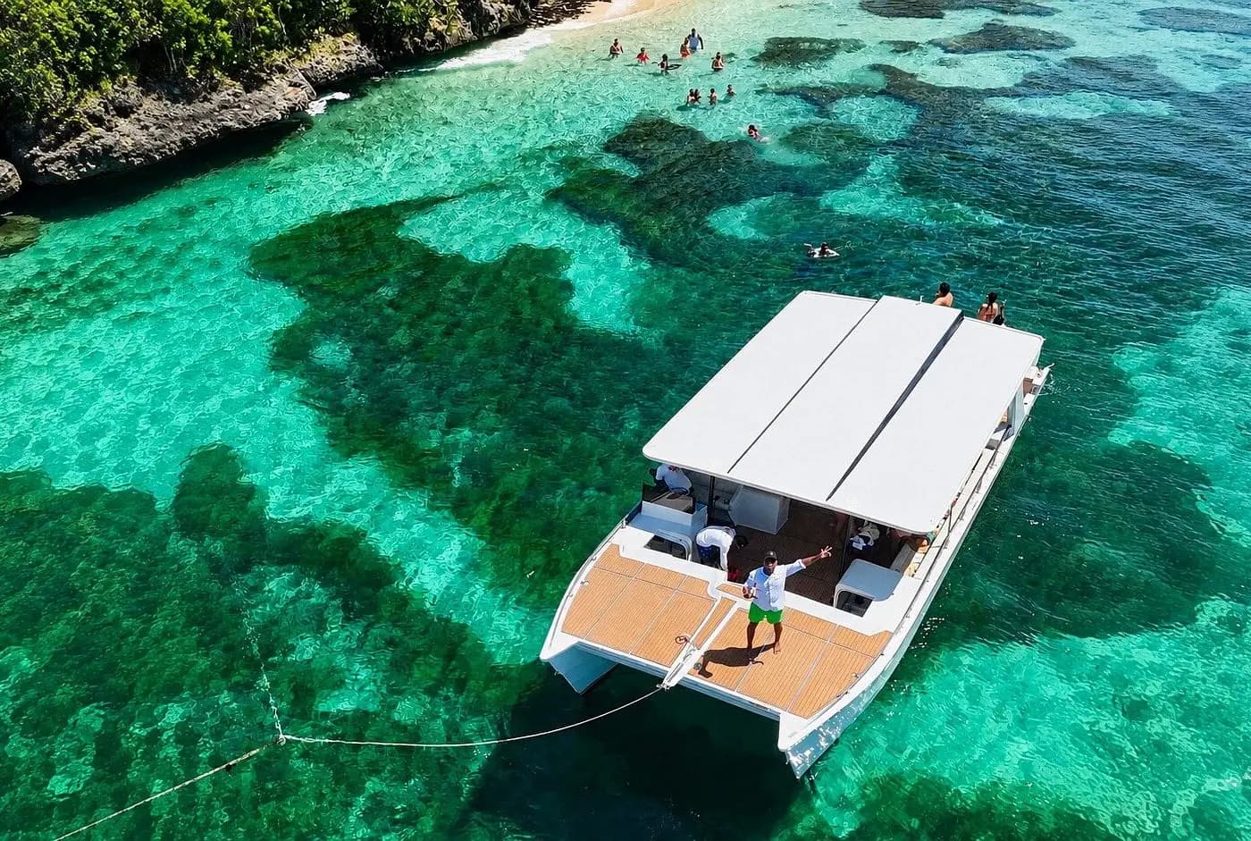 Aerial view of a power catamaran anchored over turquoise reef shallows as swimmers head for a rocky Samaná shore