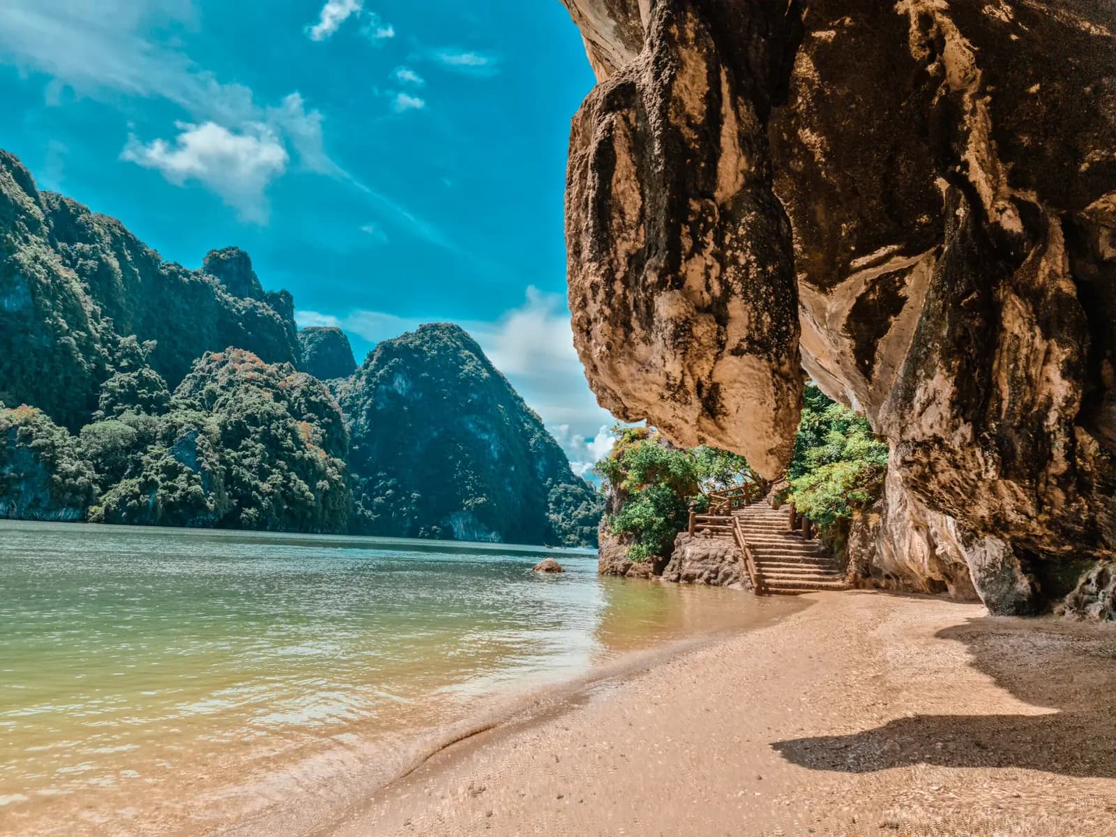 Steps climbing beneath an overhanging limestone cliff beside a calm Phang Nga Bay beach