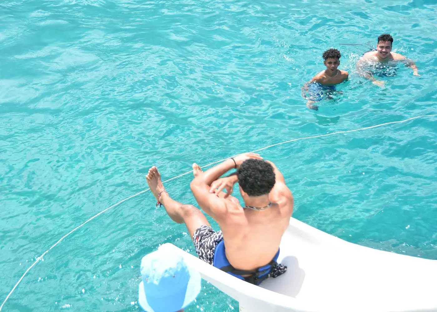Man riding the catamaran's waterslide toward two swimmers in turquoise water