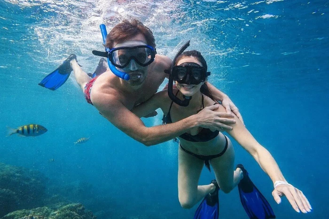 Snorkeling couple in fins embracing underwater beside a small striped fish