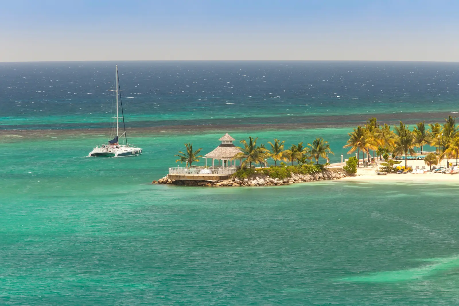 A sailing catamaran anchored off a palm-fringed point with a thatched gazebo in Ocho Rios