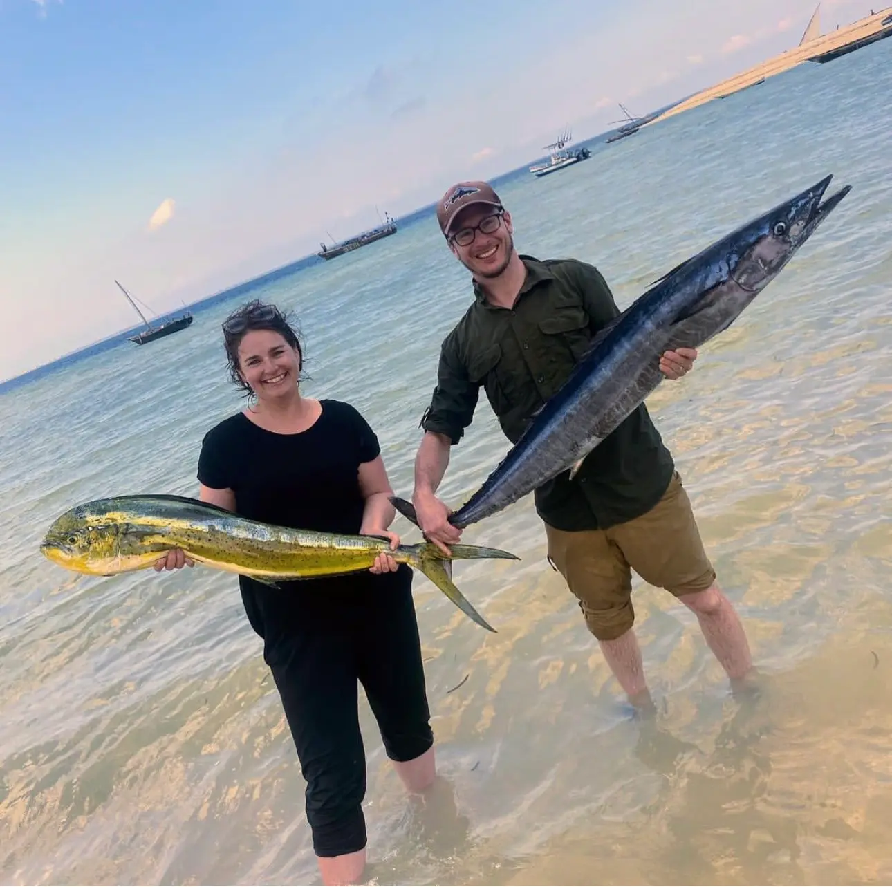 Two anglers standing in beach shallows with a mahi-mahi and a wahoo from the day's catch