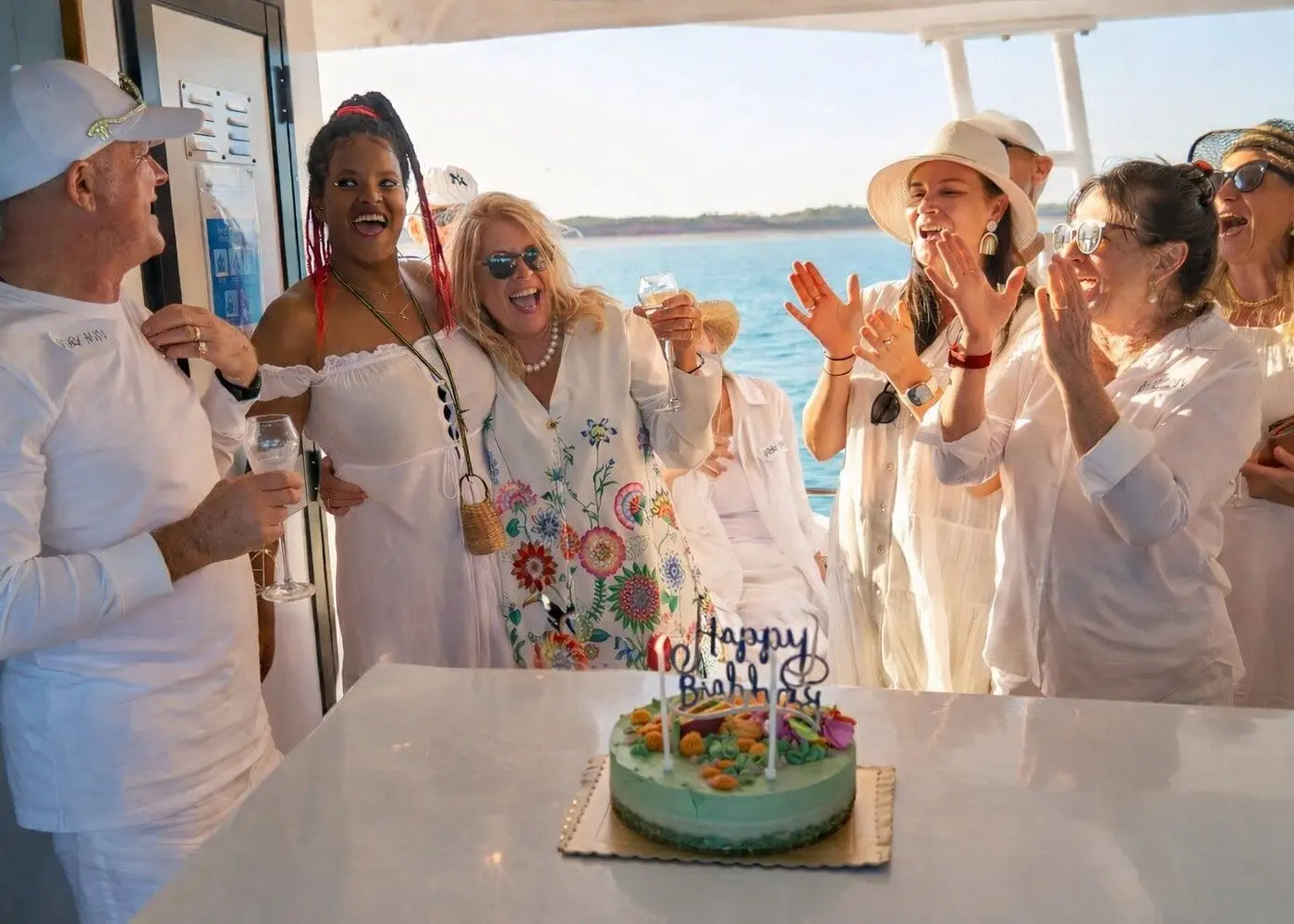 Guests dressed in white clapping around a birthday cake on the catamaran deck