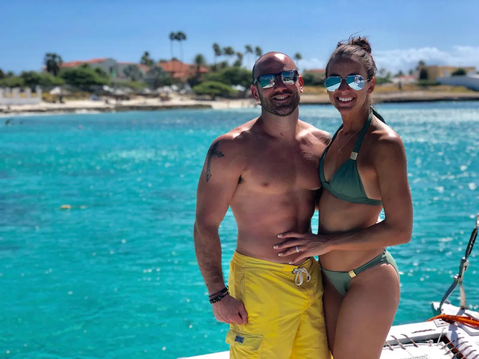 Couple posing on a catamaran deck with turquoise water and Aruba's palm-lined shore behind