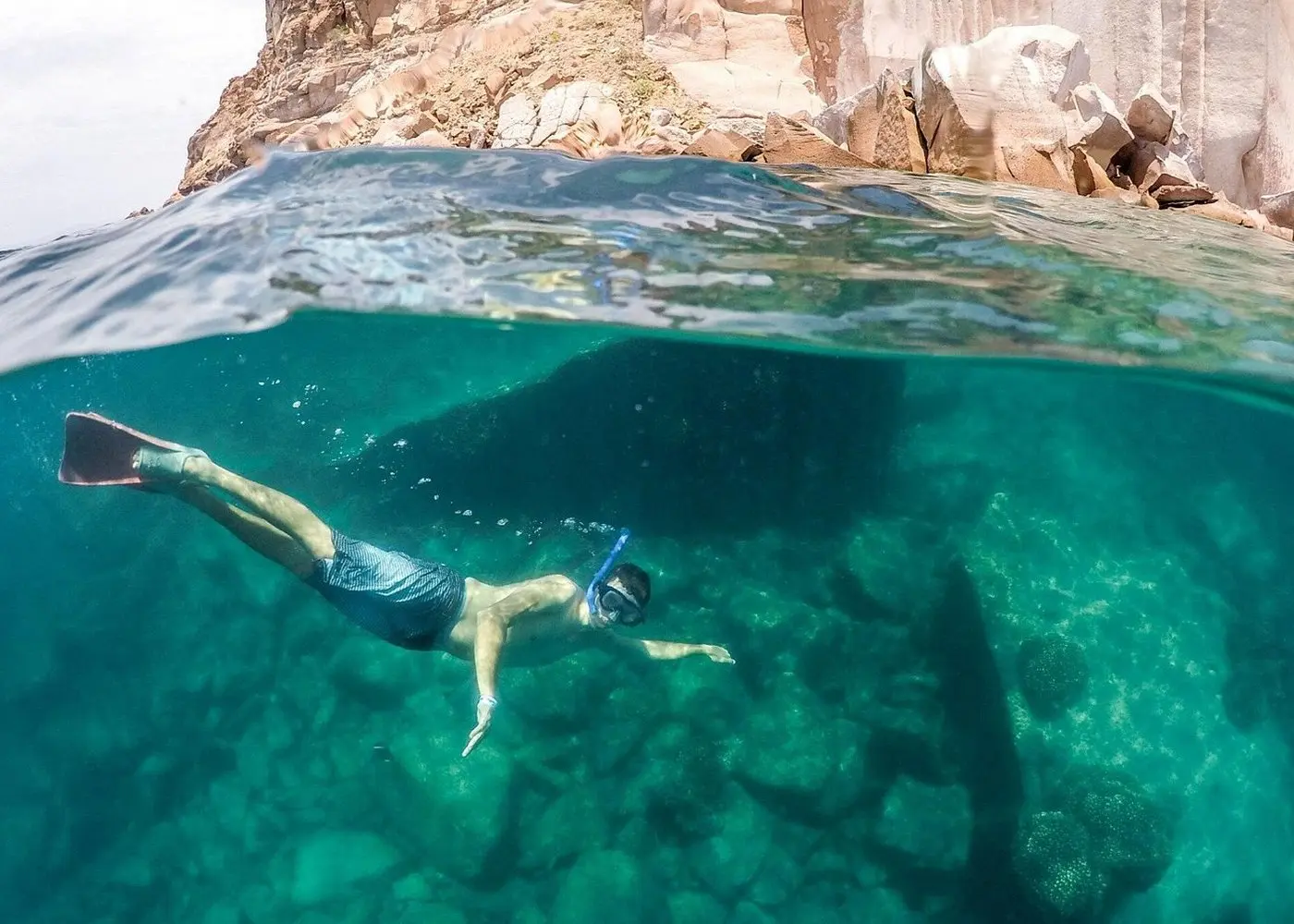 Over-under view of a snorkeler diving past a rocky reef below a red rock cliff