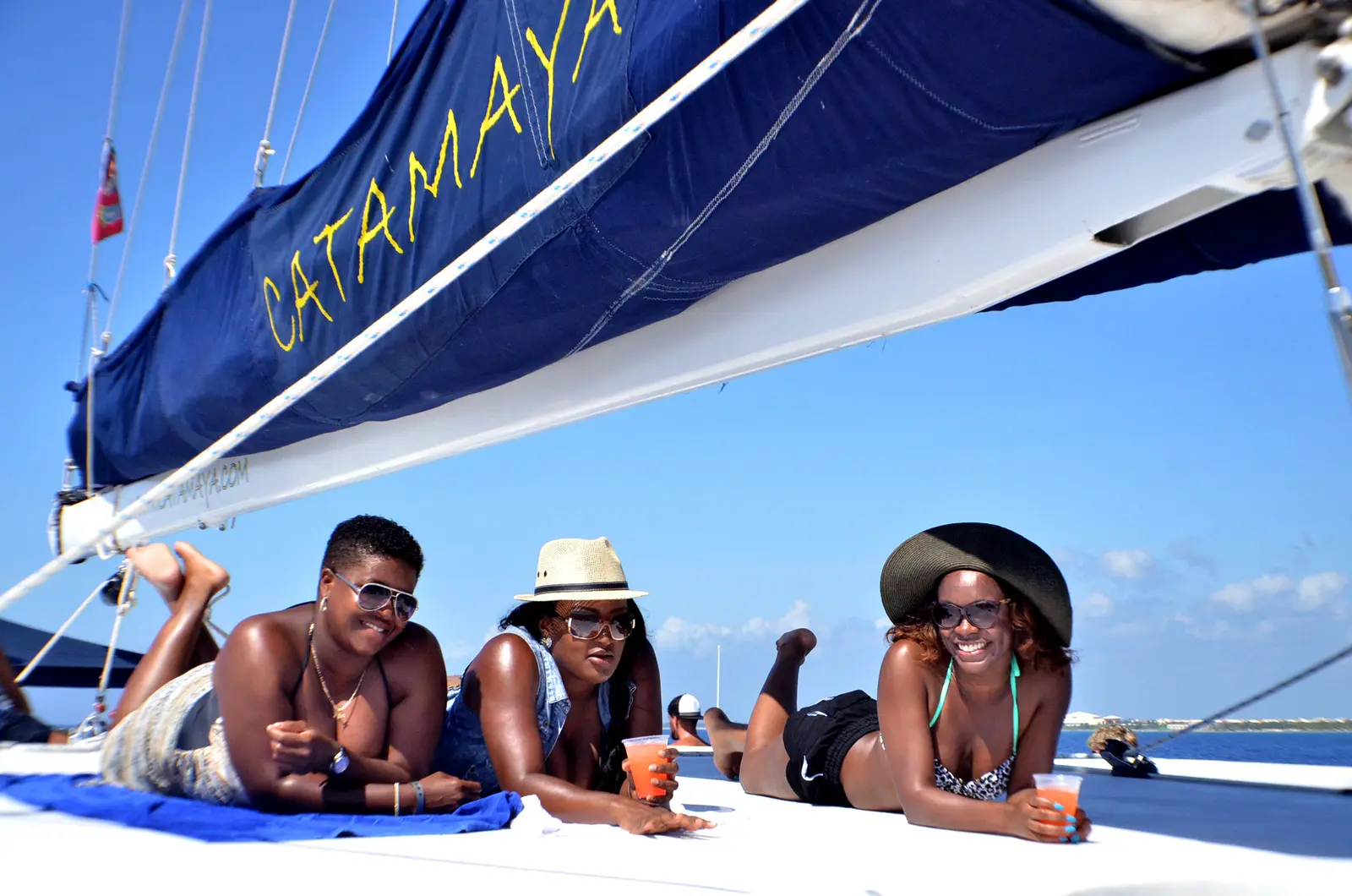 Three friends lying on the catamaran's sunny deck with drinks beneath the furled sail
