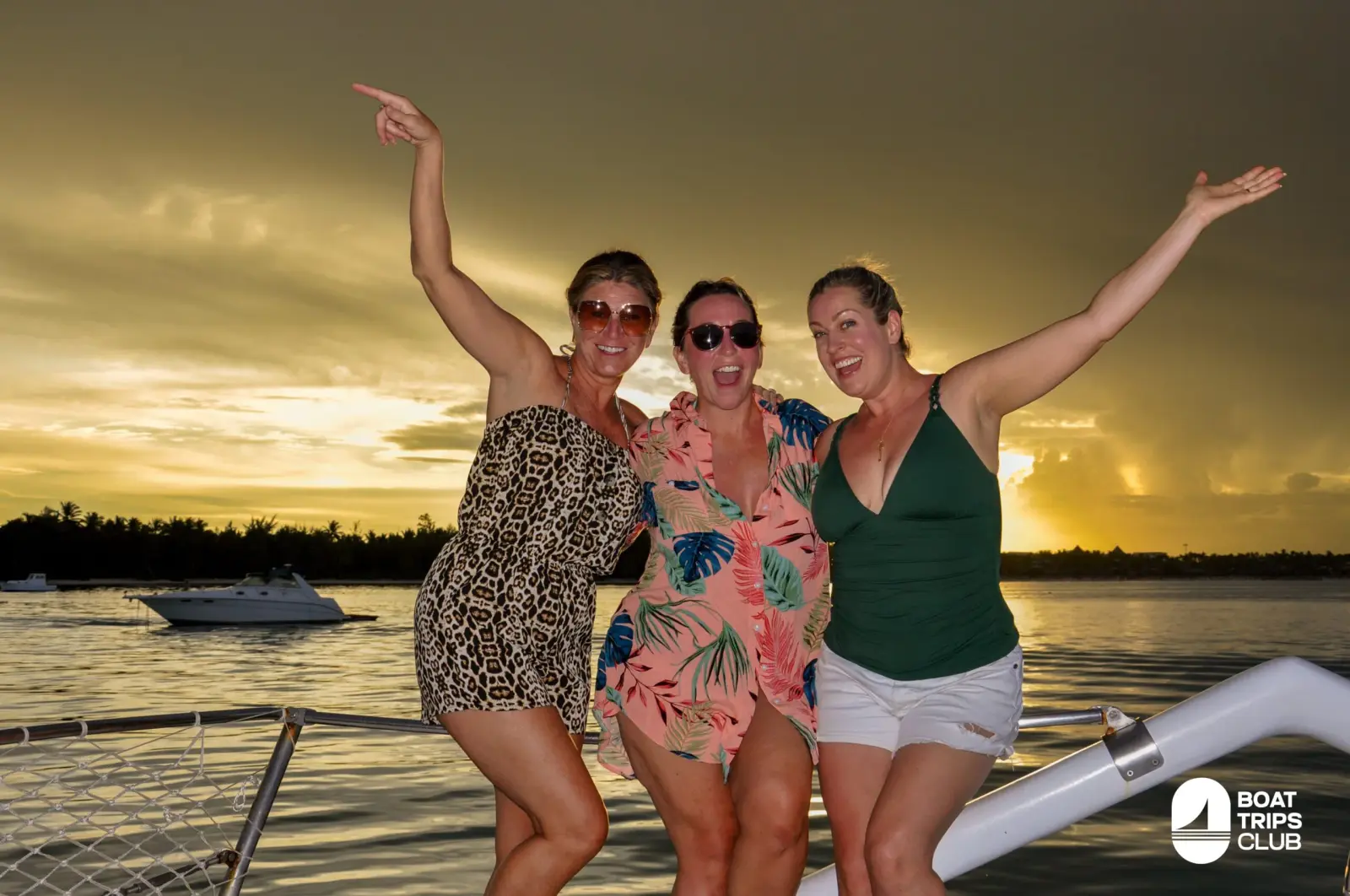 Three women throw their arms wide at the bow as a golden sunset glows over Punta Cana