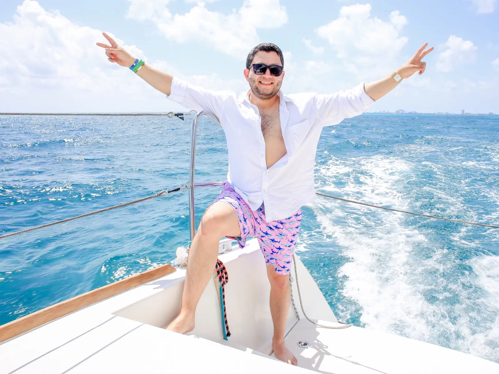 Man in a white shirt flashing peace signs on a speeding yacht's deck, Cancún skyline faint on the horizon