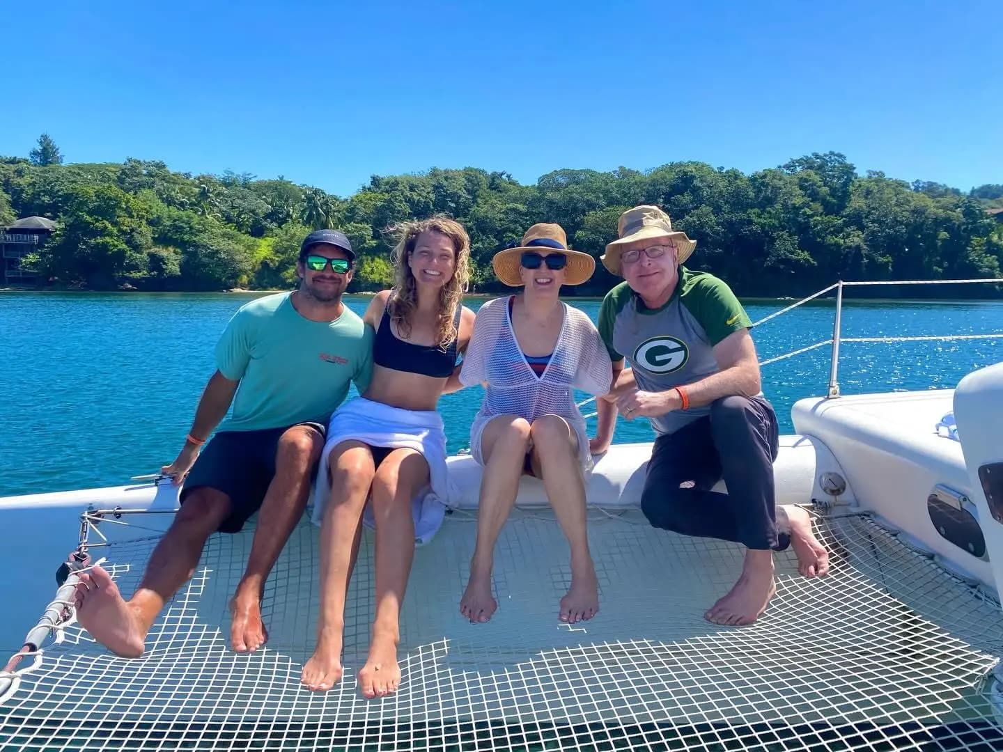 Four adults in hats and sunglasses sitting on the catamaran's trampoline, wooded Roatan shore behind