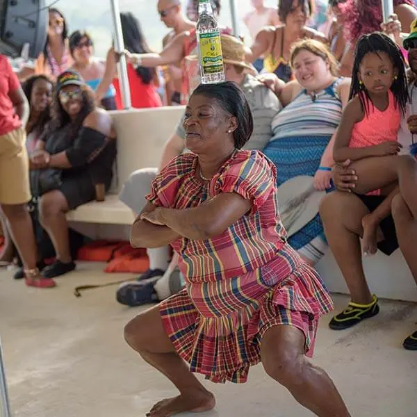 A dancer in a plaid dress squatting low with a bottle on her head as guests watch on the catamaran