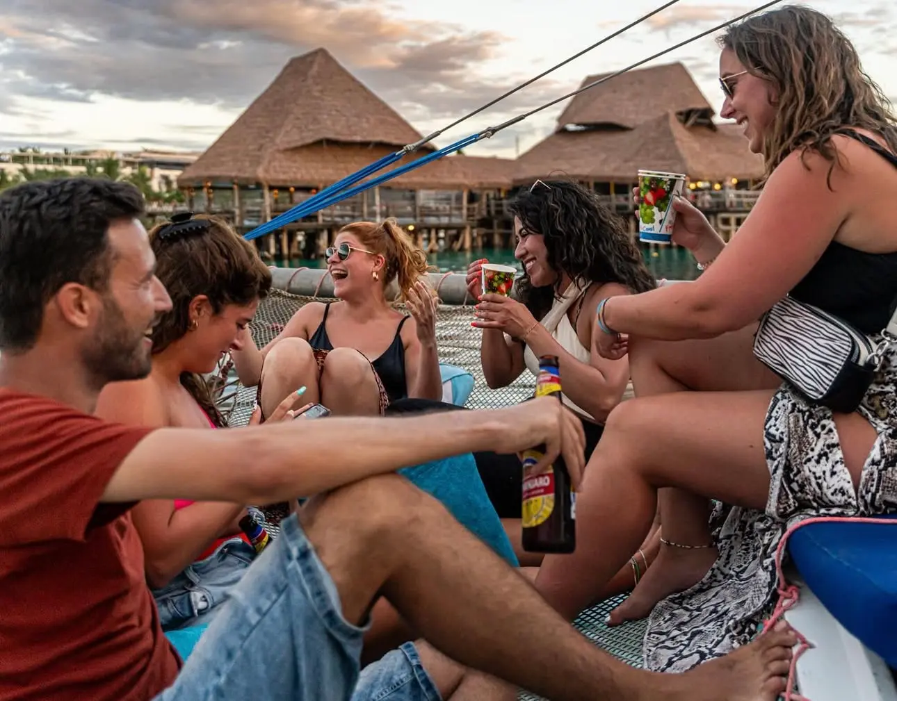 Guests laughing over drinks on a catamaran's deck, thatched-roof buildings on the Zanzibar shore behind
