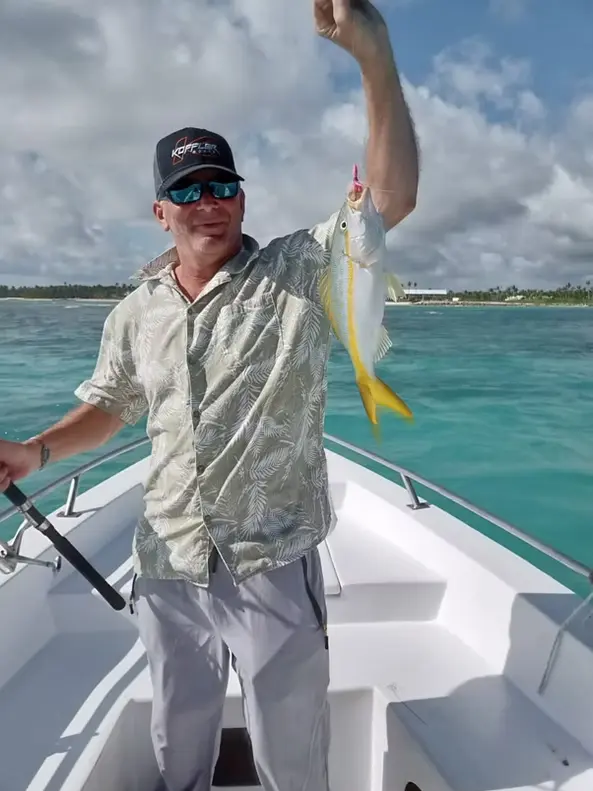 Man in a cap holding up a yellowtail snapper at the bow of a fishing boat over turquoise water