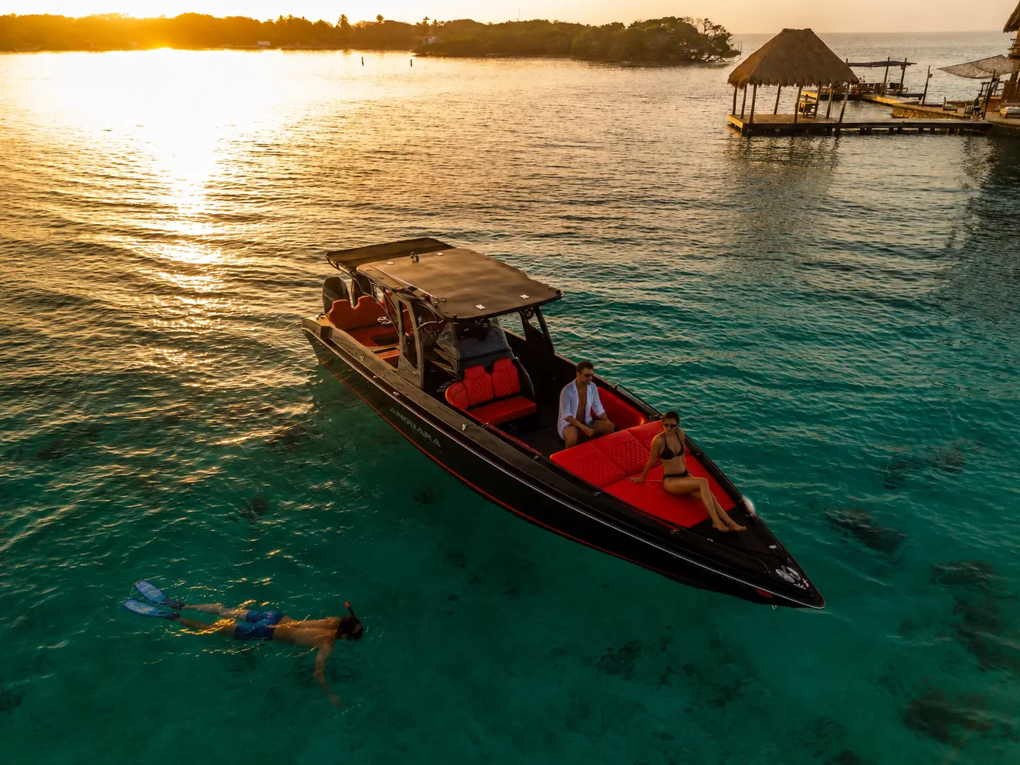 Black speedboat with red seats anchored by a thatched dock at sunset in the Rosario Islands, a snorkeler nearby