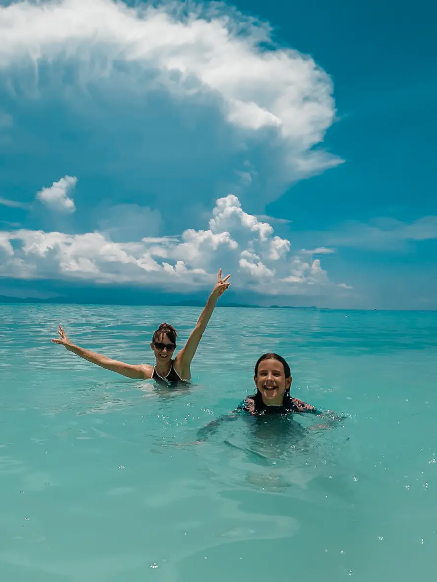 Two women waving from chest-deep turquoise water under towering white clouds off Racha Island