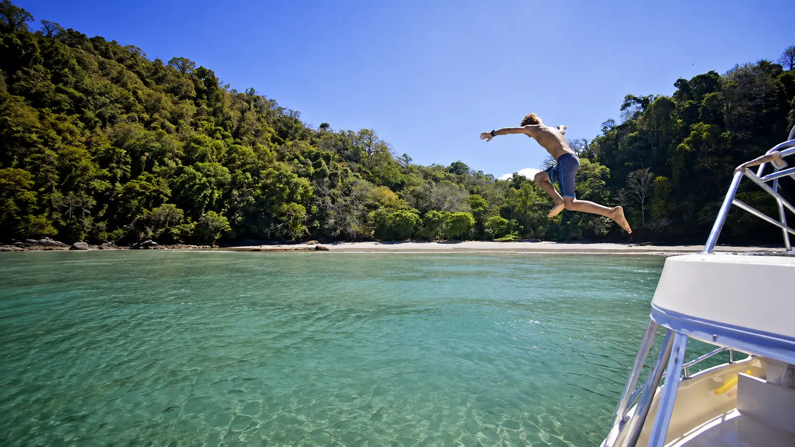 Man leaping from the catamaran into clear turquoise water off a forested Tortuga Island beach