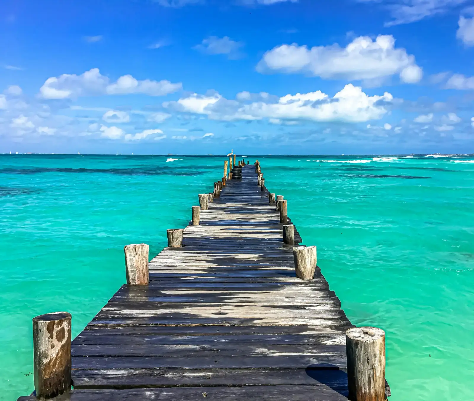 Weathered wooden pier stretching into turquoise Caribbean water under a blue sky with puffy white clouds