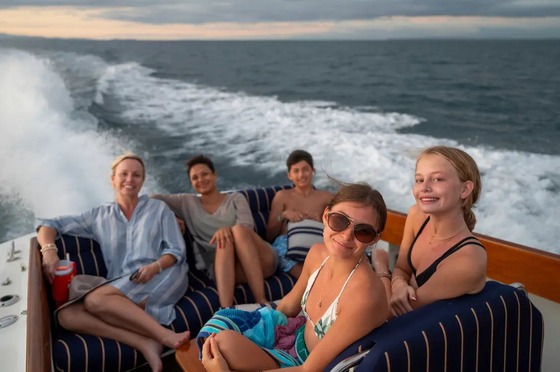 Five guests smiling on striped cushions in the stern of a picnic boat, white wake trailing behind