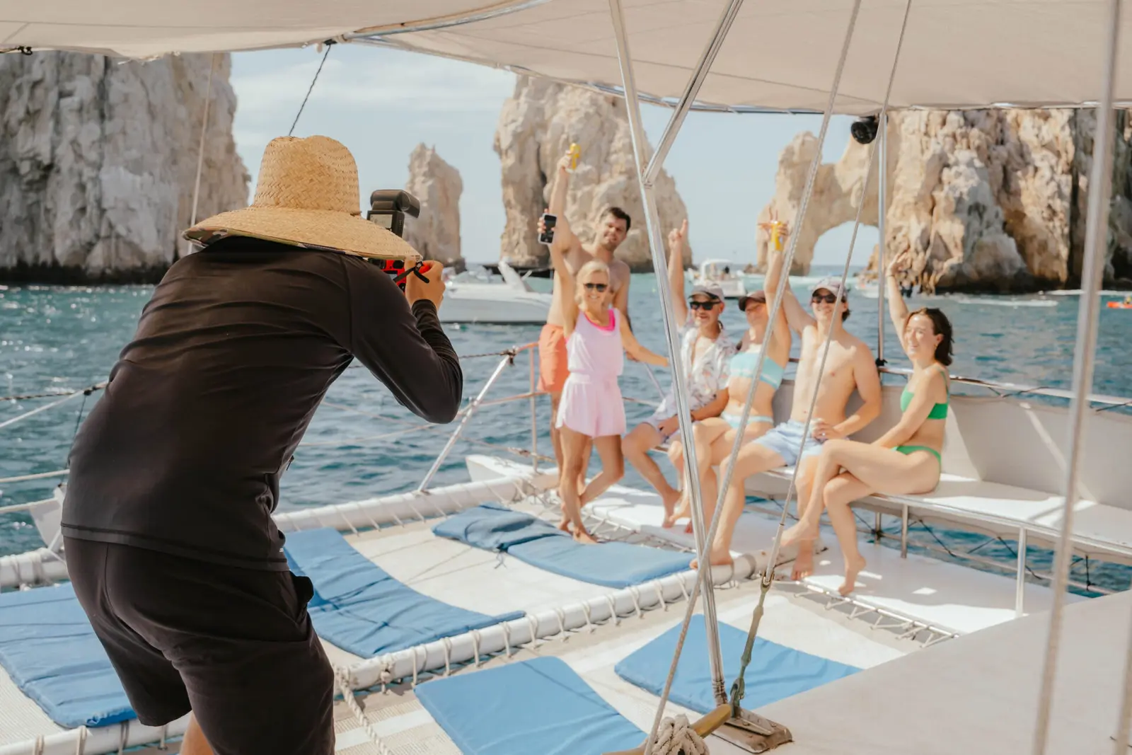 Photographer in a straw hat captures guests posing on the catamaran with the Land's End rocks behind