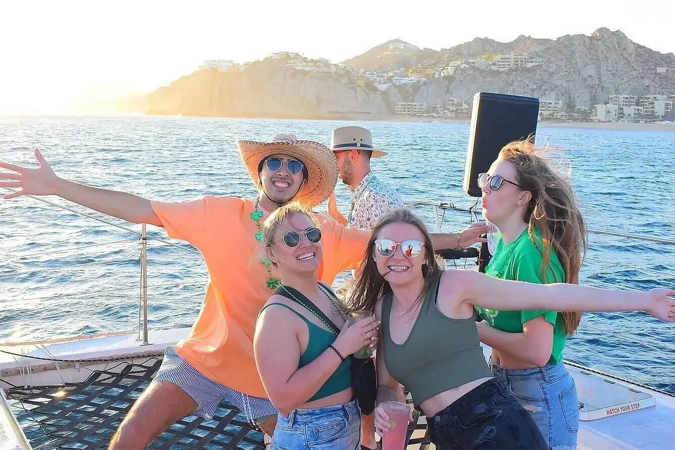 Friends striking poses on a catamaran's bow net at golden hour near a hilly coast