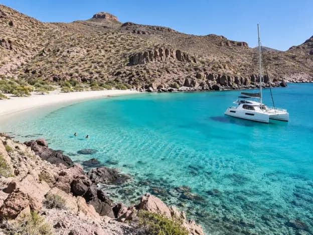 White catamaran anchored in turquoise waters near Ensenada Grande Beach on Espíritu Santo Island, with sandy shores and rocky desert hills.