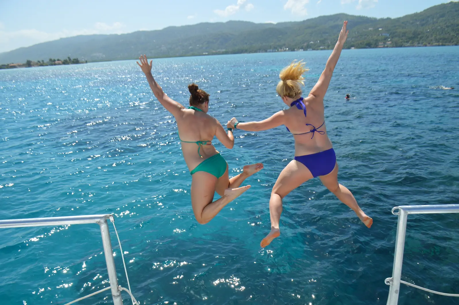 Two women jumping hand in hand off a catamaran into the sea with Montego Bay's green hills behind