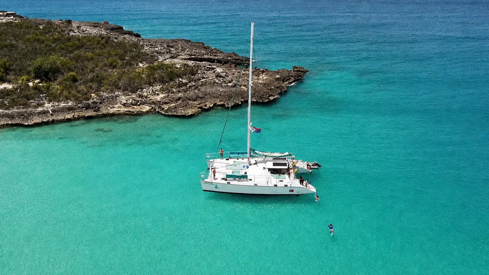 Aerial view of a sailing catamaran anchored in turquoise water off a rocky St. Maarten headland, a swimmer nearby