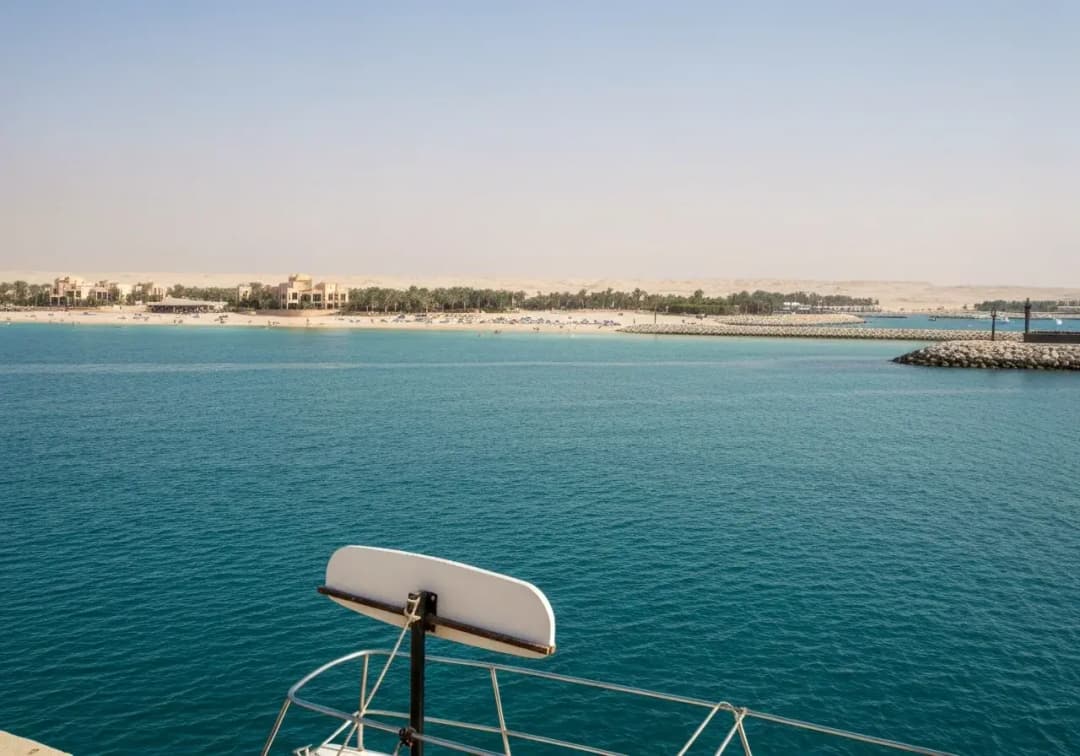 View past a boat railing across turquoise water to a palm-lined beach and rock breakwater on the Dubai coast