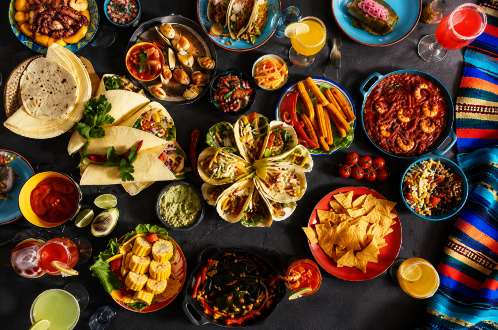 Traditional Mexican food spread with tacos, guacamole, salsa, seafood, chips, and colorful drinks.