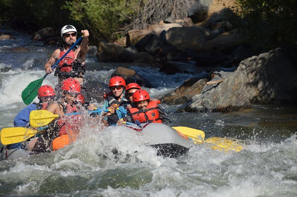 A group of people in a raft navigating through turbulent waters, wearing helmets and life jackets. The scene captures the excitement of whitewater rafting, with splashes of water around them.