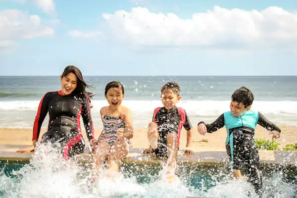A woman and three children joyfully splashing water while sitting on the edge of a pool, with a beach and ocean in the background.