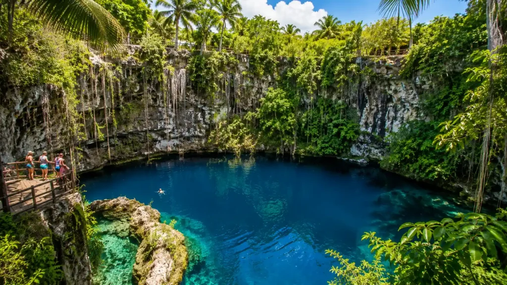 Clear blue Hoyo Azul cenote surrounded by limestone cliffs, tropical greenery, wooden viewing deck, and visitors enjoying an inland Cap Cana excursion.