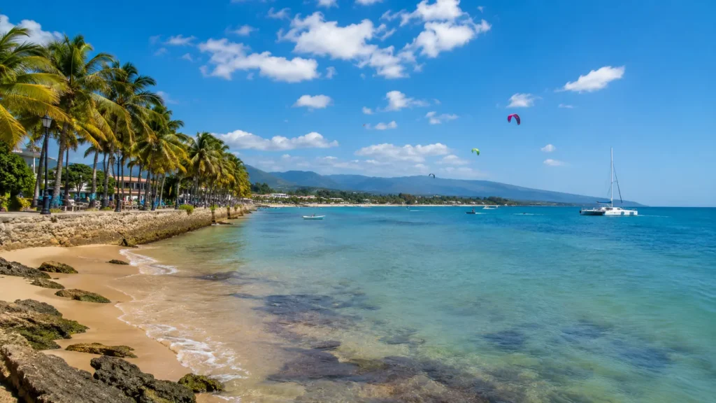 Sunny Puerto Plata coastline in March with clear turquoise water, palm-lined promenade, catamaran, kiteboarders, and mountains in the background.