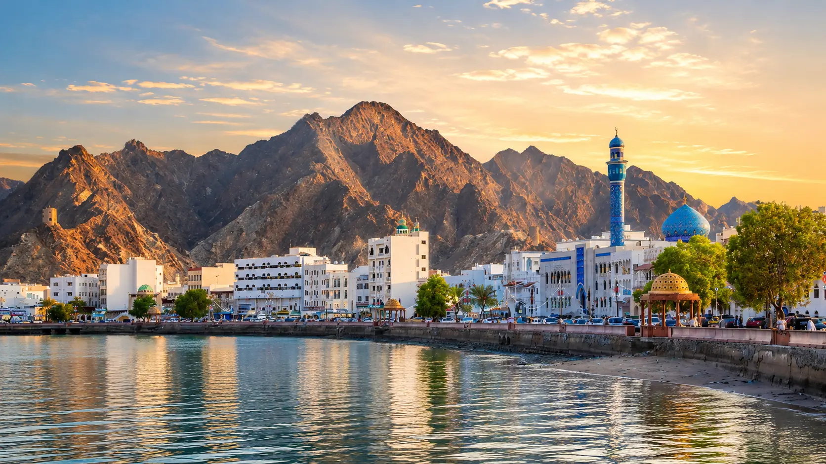 Things to do in Oman featuring Muscat's scenic waterfront, majestic Hajar Mountains, blue mosque, and calm coastline at sunrise.