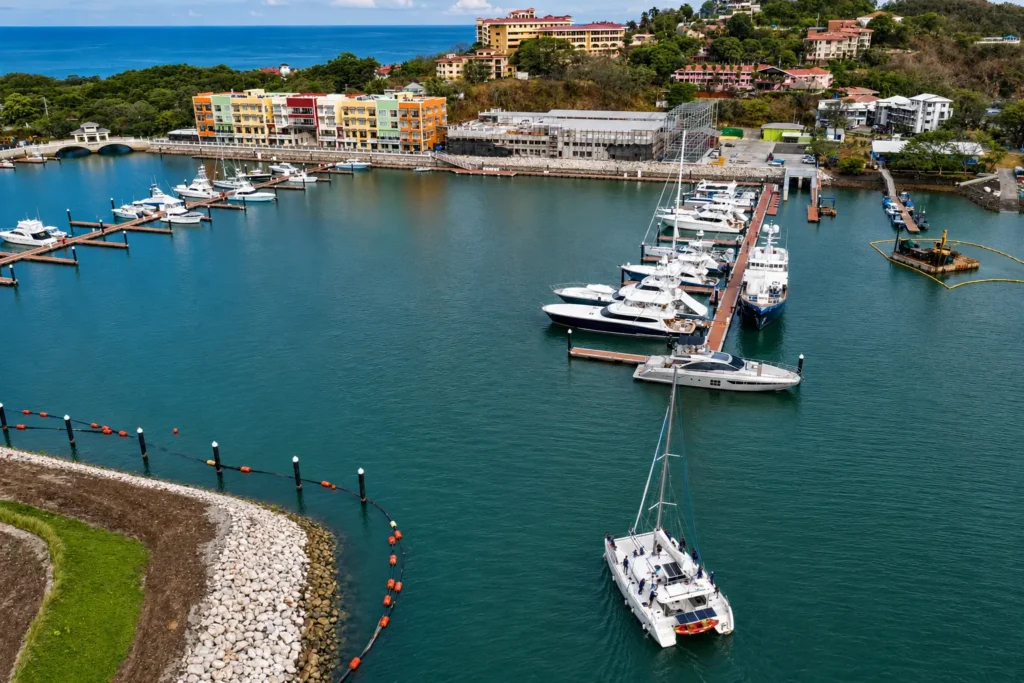 A high-resolution aerial view of a Costa Rica marina featuring luxury yachts, a sailing catamaran, waterfront buildings, and a scenic Pacific coastline surrounded by lush hills.
