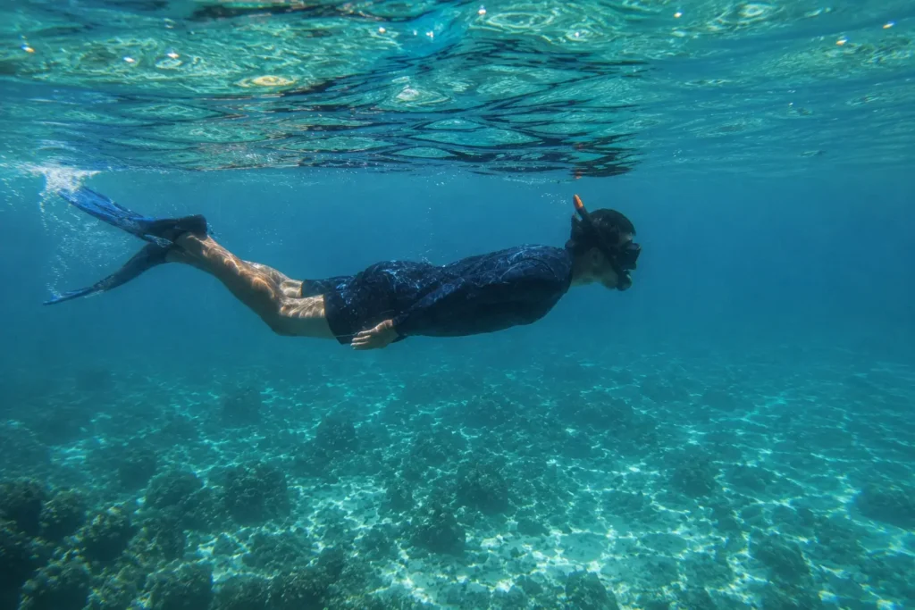 Person snorkeling underwater in Saona Island's crystal-clear turquoise sea, wearing a full-body rash guard and fins above a tropical coral reef.