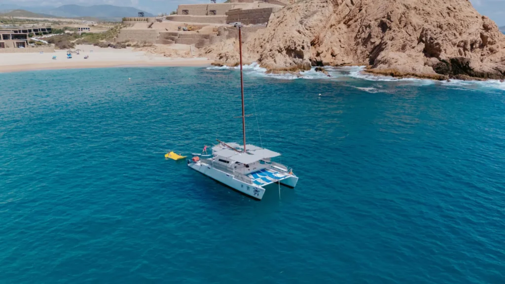 Catamaran anchored in clear turquoise water off the Cabo coastline near a secluded beach and rocky cliffs, showcasing a peaceful sailing tour in Cabo San Lucas.