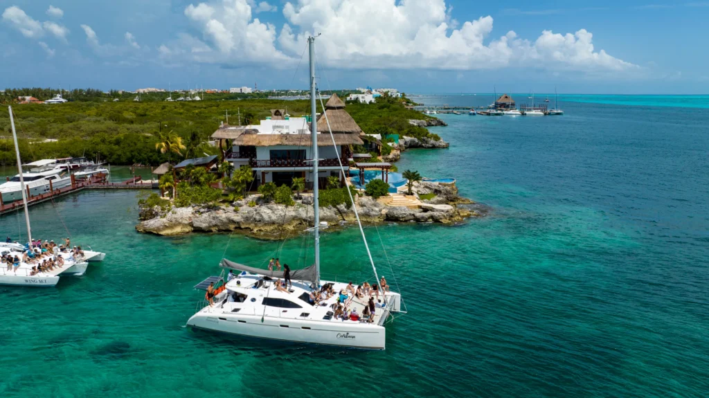 Catamaran tour sailing across the turquoise waters of Cancun near a tropical island and marina with the Caribbean coastline in the background.